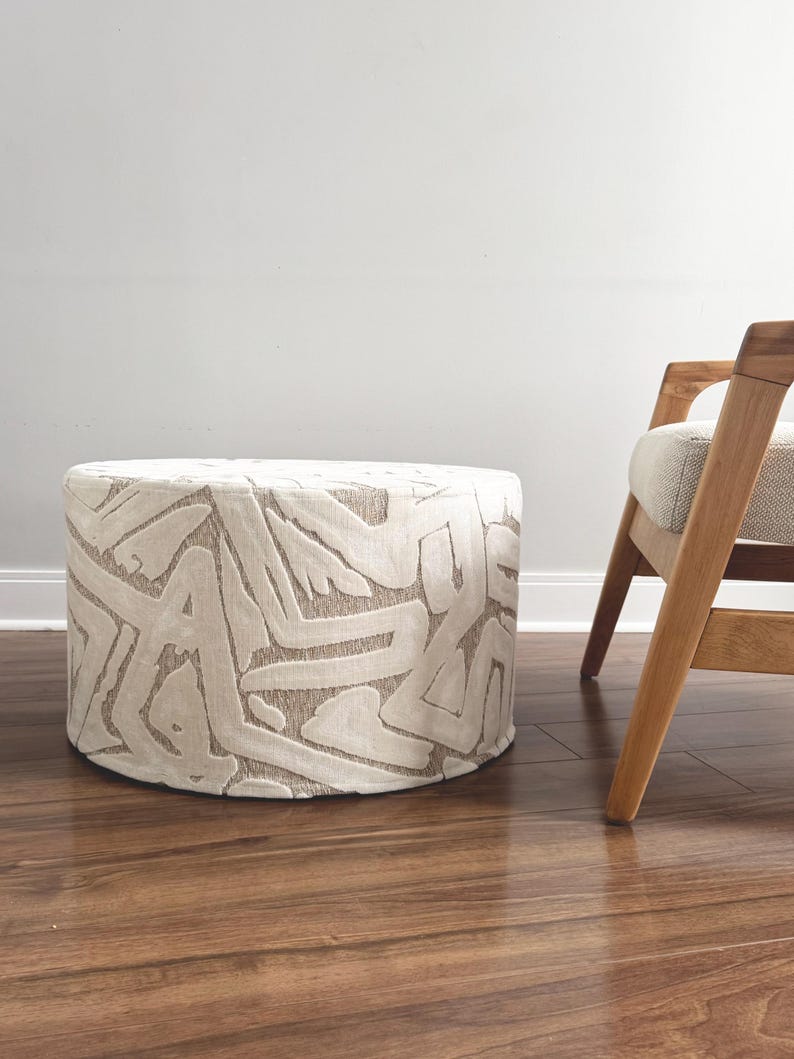 Puede incluir: Un otom&aacute;n cil&iacute;ndrico con tela color crema con un patr&oacute;n abstracto repetido en un tono marr&oacute;n claro. El otom&aacute;n est&aacute; sobre un piso de madera, con una silla de madera visible en el fondo.