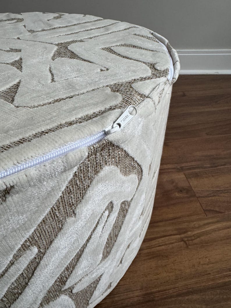 Puede incluir: Un otom&aacute;n redondo y tapizado con un dise&ntilde;o estampado crema y marr&oacute;n. La tela tiene un aspecto texturizado en relieve. Una cremallera blanca recorre el lateral. El otom&aacute;n est&aacute; sobre un suelo de madera.