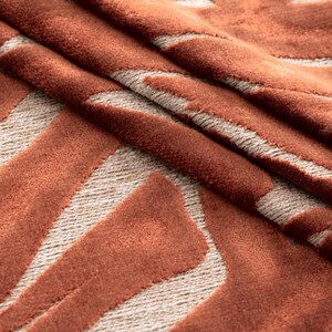 Puede incluir: Primer plano de una alfombra plegada con un patr&oacute;n geom&eacute;trico. La alfombra presenta un dise&ntilde;o texturizado de color &oacute;xido sobre un fondo beige. El patr&oacute;n consiste en formas angulares, creando una est&eacute;tica moderna. Los pliegues de la alfombra a&ntilde;aden profundidad.