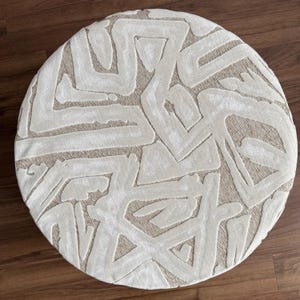 Puede incluir: Un otom&aacute;n redondo beige con un patr&oacute;n geom&eacute;trico abstracto crema. El dise&ntilde;o presenta l&iacute;neas gruesas y texturizadas que crean un aspecto moderno y art&iacute;stico. El otom&aacute;n est&aacute; colocado sobre un suelo de madera.