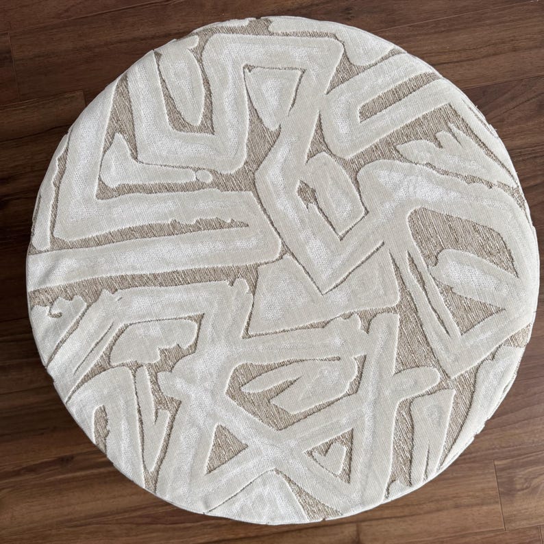 Puede incluir: Un otom&aacute;n redondo beige con un patr&oacute;n geom&eacute;trico abstracto crema. El dise&ntilde;o presenta l&iacute;neas gruesas y texturizadas que crean un aspecto moderno y art&iacute;stico. El otom&aacute;n est&aacute; colocado sobre un suelo de madera.