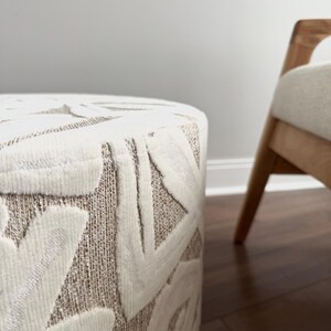 Puede incluir: Un otom&aacute;n cil&iacute;ndrico con una tela texturizada de color crema con un patr&oacute;n geom&eacute;trico repetitivo en un tono beige claro. Una silla de madera con un coj&iacute;n de color claro es parcialmente visible en el fondo.