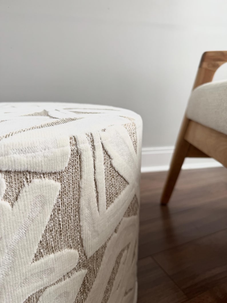 Puede incluir: Un otom&aacute;n cil&iacute;ndrico con una tela texturizada de color crema con un patr&oacute;n geom&eacute;trico repetitivo en un tono beige claro. Una silla de madera con un coj&iacute;n de color claro es parcialmente visible en el fondo.