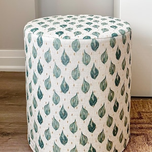May include: A cylindrical ottoman upholstered in cream fabric, patterned with green leaf motifs and gold dots. The footstool is on a brown rug. The height is approximately 40 cm.