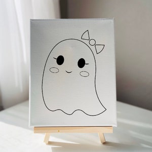May include: A white canvas with a black line drawing of a ghost with a bow on its head. The ghost has large eyes and a small smile. The canvas is on a small wooden easel.