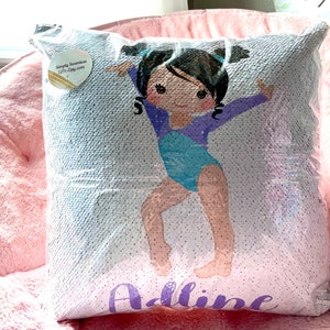 May include: A white and purple sequin pillow with a cartoon image of a girl in a blue leotard doing a gymnastics pose. The pillow has the name "Addiline" written in purple sequins.