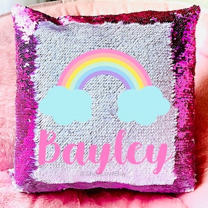 May include: A pink and white sequin pillow with a rainbow and clouds design. The pillow has the name "Bailey" written on it in pink.