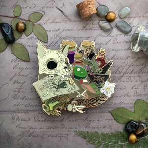 May include: A gold-toned enamel pin depicting a cat skull, a frog, a potion bottle, and a book with bones and herbs. The pin is set on a wooden table with a white flower and a green potion bottle.