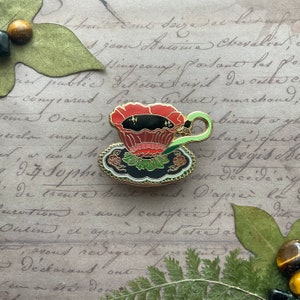 May include: A black and gold enamel pin of a teacup with a red flower design. The teacup is filled with black tea and has a green handle. The teacup is sitting on a saucer with a black and gold floral design.