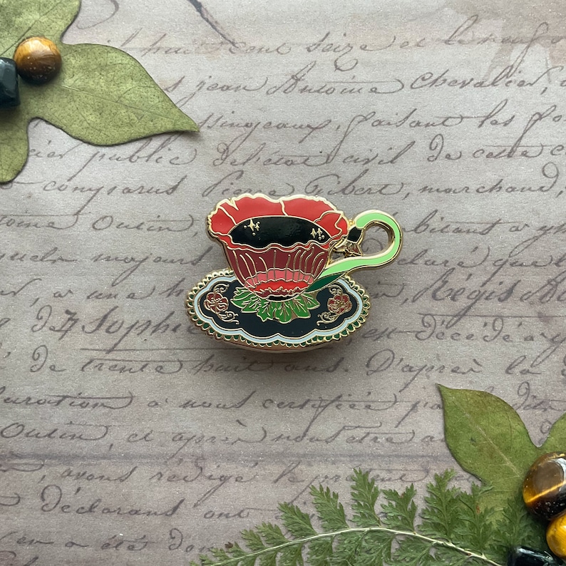 May include: A black and gold enamel pin of a teacup with a red flower design. The teacup is filled with black tea and has a green handle. The teacup is sitting on a saucer with a black and gold floral design.