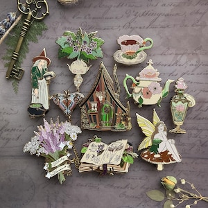 May include: A collection of ten enamel pins featuring whimsical designs, including a witch, a fairy, a teapot, a book, a house, a flower, a butterfly, and a mandrake. The pins are gold-toned and have a variety of colors.