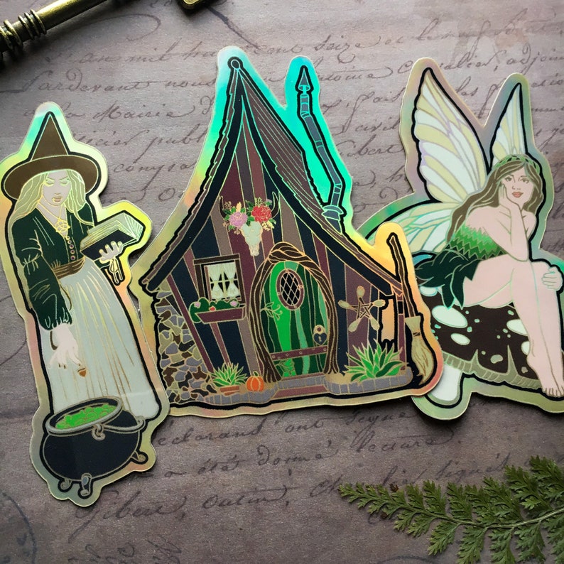 May include: Three iridescent stickers featuring a witch holding a book, a cottage with a green door, and a fairy with green wings. The stickers are on a brown background with writing.
