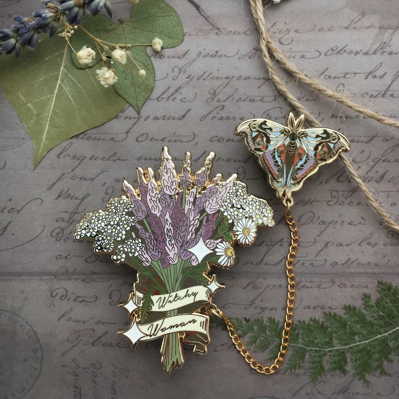 May include: Two gold-tone enamel pins with a chain connecting them. The first pin features a bouquet of lavender, white flowers, and greenery with a banner that reads "Witchy Woman". The second pin features a butterfly with pink, blue, and orange wings.