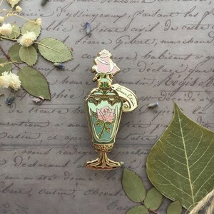 May include: Gold-tone enamel pin of a vintage glass bottle with a rose inside. The bottle has a gold stopper and a tag that reads "Doux Elixir".