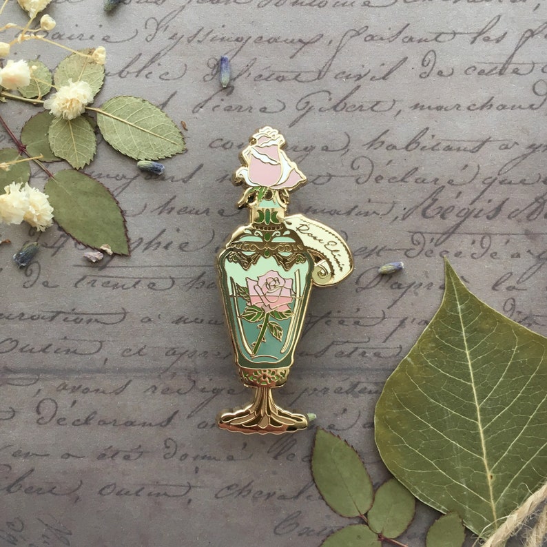May include: Gold-tone enamel pin of a vintage glass bottle with a rose inside. The bottle has a gold stopper and a tag that reads "Doux Elixir".
