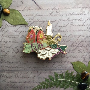 May include: A gold-toned enamel pin depicting a bundle of sage, a candle, and a cauldron with runes. The pin is set against a background of old paper with handwritten text.