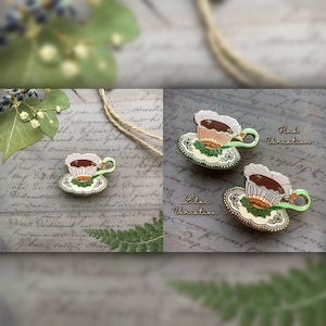 May include: Two enamel pins featuring a teacup and saucer with a floral design. The teacups are filled with brown liquid and have a white, pink, or lilac colored rim. The saucers are green and gold with a floral design. The text "Pink Variation" and "Lilac Variation" is written below the pins.