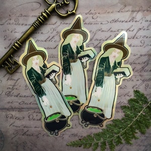 May include: Three iridescent gold stickers featuring a witch holding a book and a cauldron. The witch is wearing a green dress and a pointed hat. The background is a vintage piece of paper with writing.