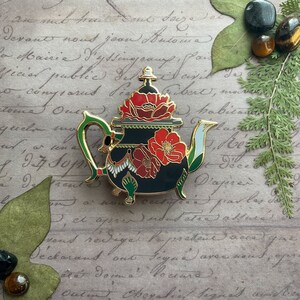 May include: A gold-tone teapot enamel pin with a black base and red poppy flowers. The teapot has a green handle and spout, and a white and gold trim.