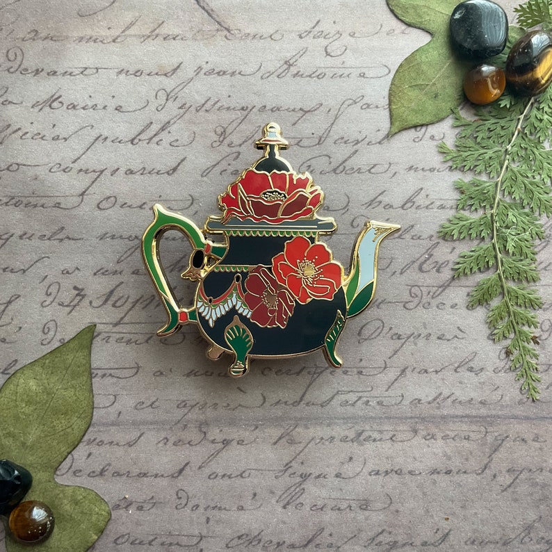 May include: A gold-tone teapot enamel pin with a black base and red poppy flowers. The teapot has a green handle and spout, and a white and gold trim.