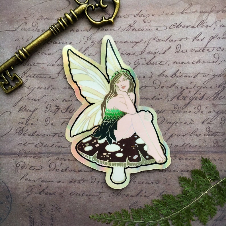 May include: A holographic sticker featuring a fairy with iridescent wings sitting on a mushroom with white spots. The fairy is wearing a green dress and has long brown hair. The background is a vintage piece of paper with writing.