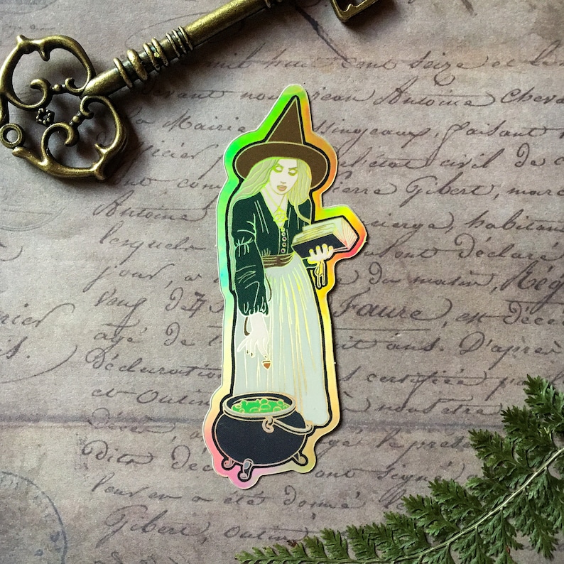 May include: A holographic sticker featuring a witch with a pointed hat, holding a book and stirring a cauldron with a green liquid. The sticker has a rainbow iridescent finish.