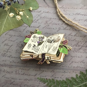 May include: A gold-toned enamel pin depicting an open book with a purple ribbon bookmark. The book is adorned with illustrations of flowers, a butterfly, and a magical symbol. The pin is surrounded by green leaves.