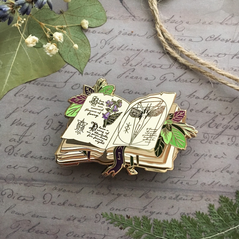 May include: A gold-toned enamel pin depicting an open book with a purple ribbon bookmark. The book is adorned with illustrations of flowers, a butterfly, and a magical symbol. The pin is surrounded by green leaves.