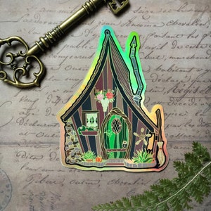 May include: A holographic sticker of a whimsical cottage with a green door, a stone foundation, and a witch's broom. The cottage has a pointed roof and a chimney. The sticker has a rainbow iridescent finish.