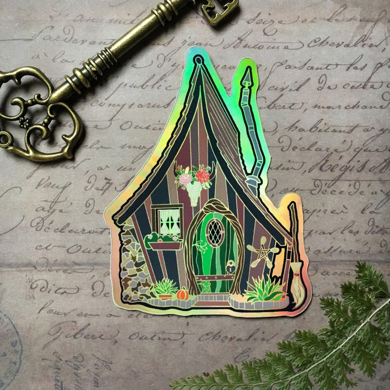 May include: A holographic sticker of a whimsical cottage with a green door, a stone foundation, and a witch's broom. The cottage has a pointed roof and a chimney. The sticker has a rainbow iridescent finish.