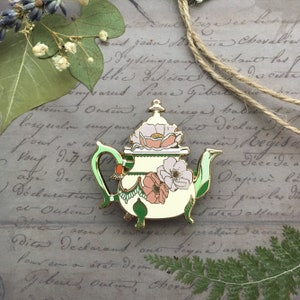 May include: A gold-tone teapot enamel pin with a green handle and floral details. The teapot is decorated with pink and white flowers and green leaves. The pin is on a background of old paper with handwritten text.