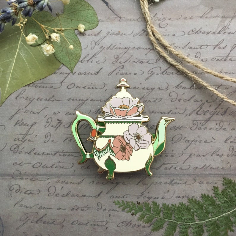 May include: A gold-tone teapot enamel pin with a green handle and floral details. The teapot is decorated with pink and white flowers and green leaves. The pin is on a background of old paper with handwritten text.