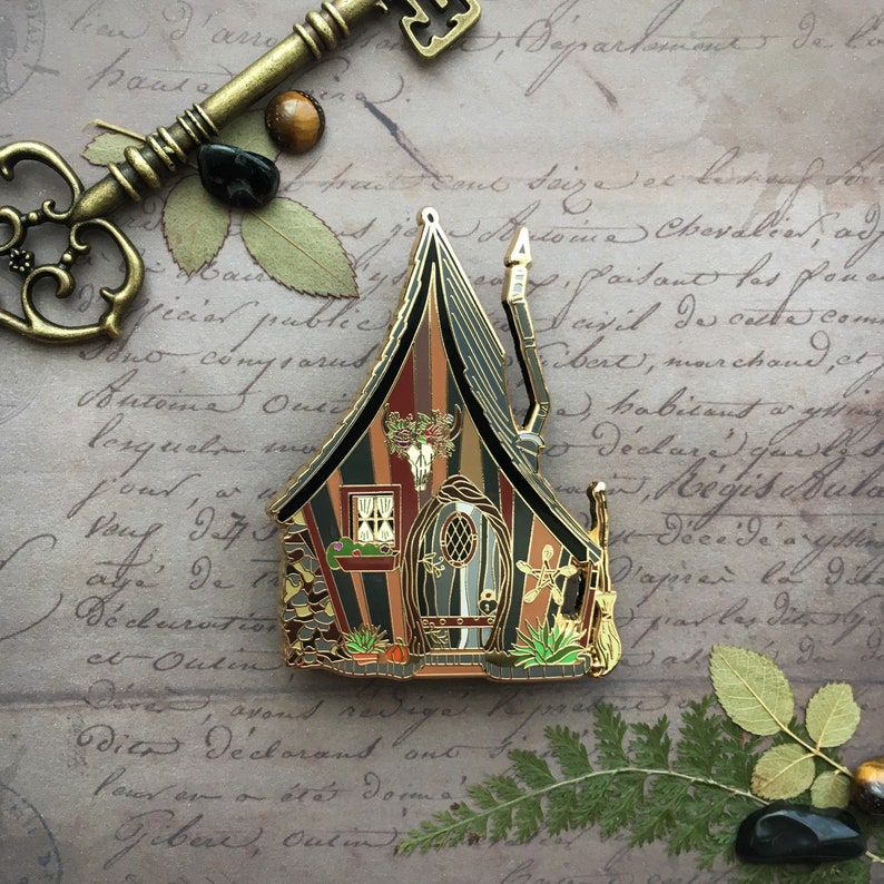 May include: A gold-toned enamel pin depicting a whimsical, striped, and detailed cottage with a pointed roof, a skull and crossbones, and a door with a crescent moon and star design.