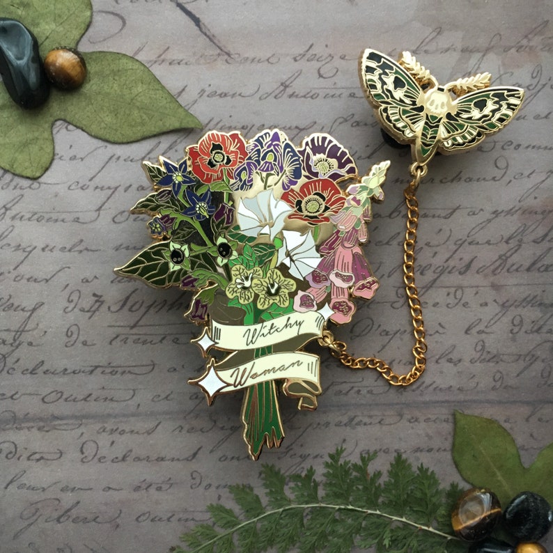 May include: Gold-tone enamel pin featuring a bouquet of flowers with a banner that reads "Witchy Woman". A gold chain connects the bouquet to a separate pin of a green and gold moth.