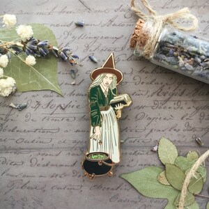 May include: A gold-toned enamel pin depicting a witch with a green dress, white skirt, and a pointed hat. She is holding a book and a cauldron with green liquid.