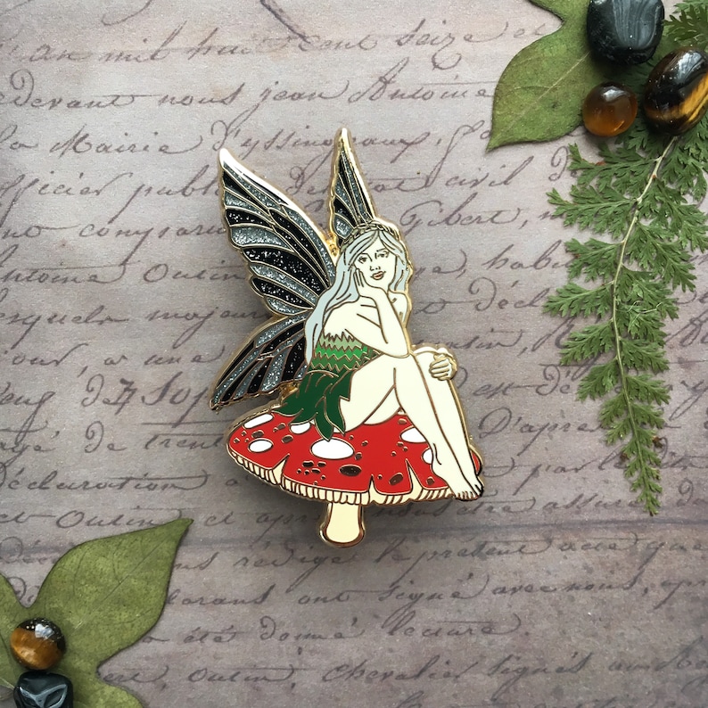 May include: A gold-toned enamel pin featuring a fairy sitting on a red and white mushroom. The fairy has silver glitter wings, long white hair, and a green dress.