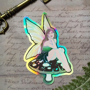 May include: A holographic sticker featuring a fairy with iridescent wings sitting on a mushroom. The fairy has long brown hair and is wearing a green dress. The mushroom is black and white with white spots.