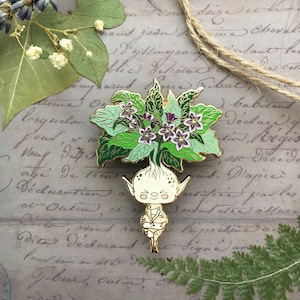 May include: A gold-tone enamel pin featuring a whimsical, smiling, humanoid figure with a green leafy crown and purple flowers. The figure is wearing a gold-tone tunic and has a long, thin body.