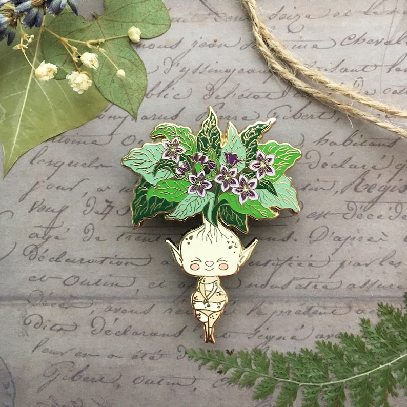 May include: A gold-tone enamel pin featuring a whimsical, smiling, humanoid figure with a green leafy crown and purple flowers. The figure is wearing a gold-tone tunic and has a long, thin body.
