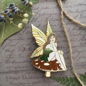 May include: Gold-tone enamel pin featuring a fairy with yellow wings sitting on a brown mushroom. The fairy has long brown hair, a green dress, and a white face.