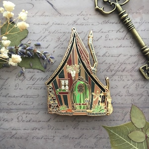 May include: A gold-tone enamel pin depicting a whimsical, multi-colored, striped cottage with a green door and a skull and horns above the door. The cottage has a pointed roof and a chimney. The pin is set against a background of old, handwritten paper.
