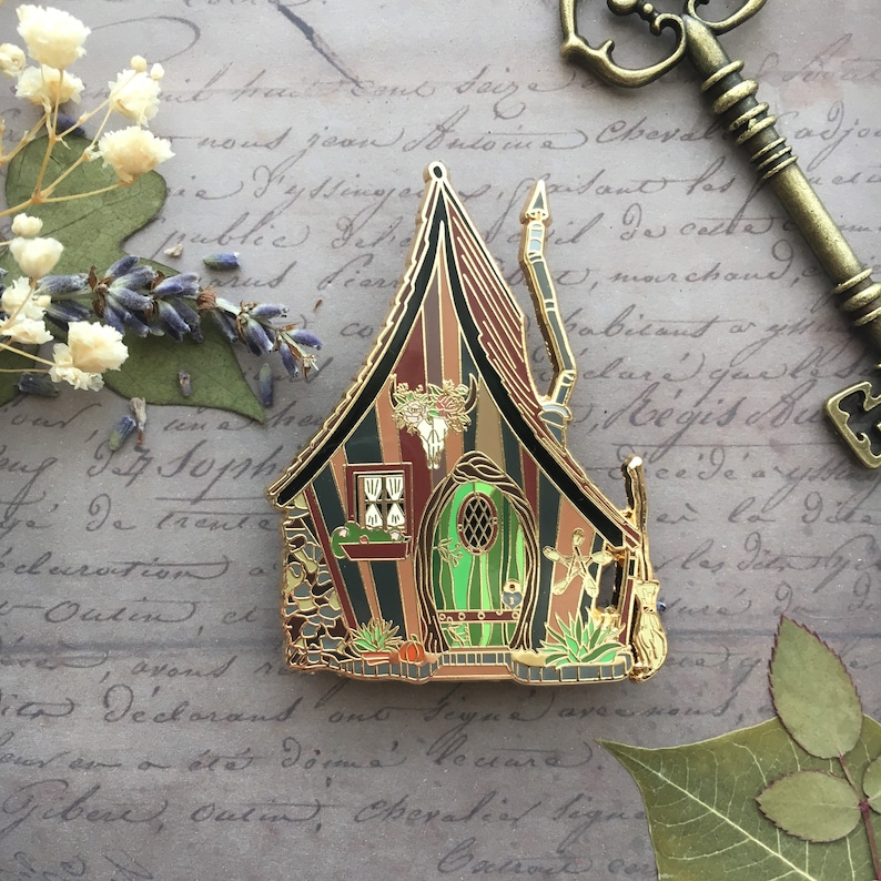 May include: A gold-tone enamel pin depicting a whimsical, multi-colored, striped cottage with a green door and a skull and horns above the door. The cottage has a pointed roof and a chimney. The pin is set against a background of old, handwritten paper.