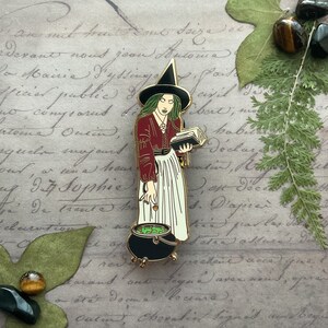 May include: A gold-toned enamel pin depicting a witch with long black hair, wearing a red dress and a black pointed hat. She is holding a book and a cauldron with green liquid. The pin is set against a background of handwritten text.
