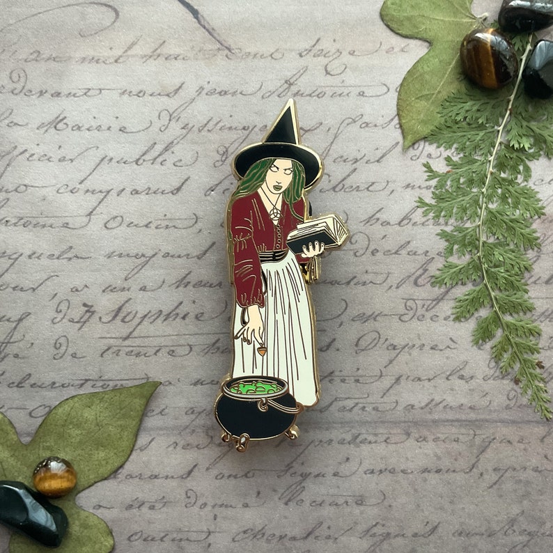 May include: A gold-toned enamel pin depicting a witch with long black hair, wearing a red dress and a black pointed hat. She is holding a book and a cauldron with green liquid. The pin is set against a background of handwritten text.
