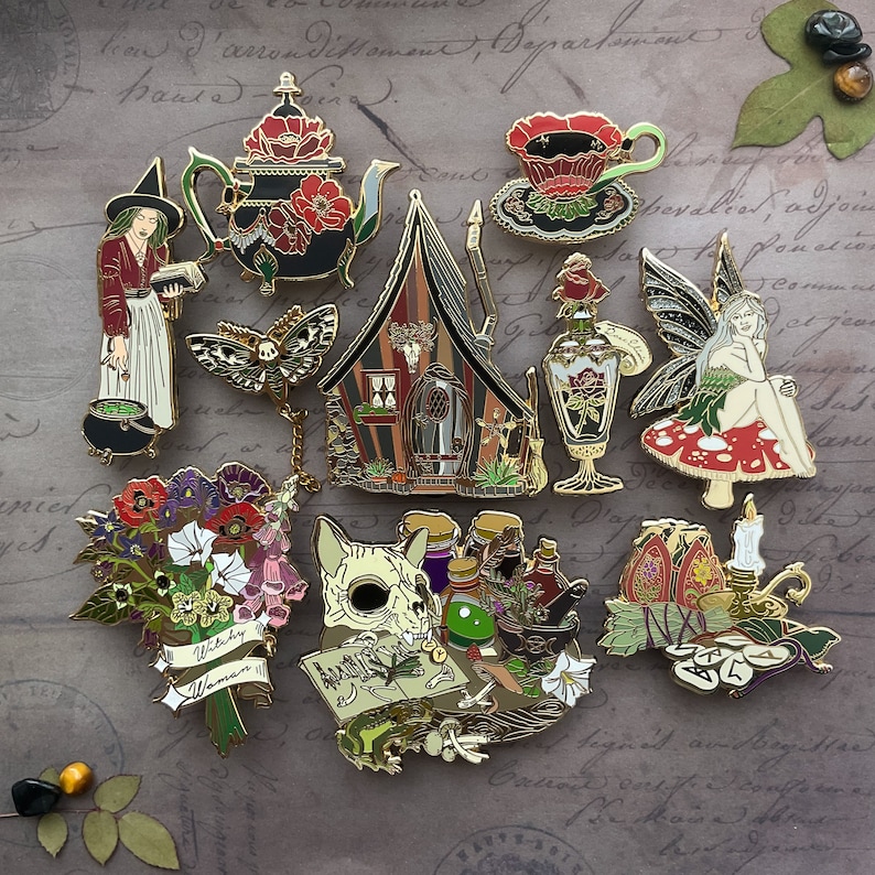 May include: A collection of enamel pins featuring a witch, a house, a potion bottle, a skull, a bouquet of flowers, a teacup, a fairy, and a candle. The pins are all gold-toned and have intricate details.