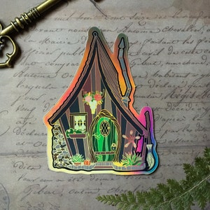 May include: A holographic sticker of a whimsical cottage with a green door, a skull and flowers above the door, and a broom and a star next to the door. The cottage is surrounded by plants and pumpkins.