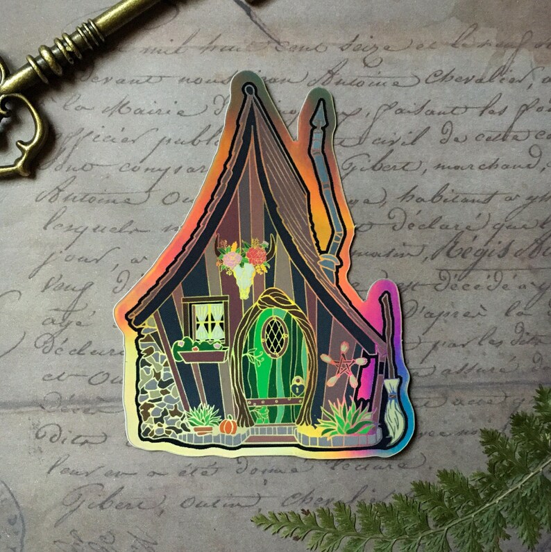 May include: A holographic sticker of a whimsical cottage with a green door, a skull and flowers above the door, and a broom and a star next to the door. The cottage is surrounded by plants and pumpkins.