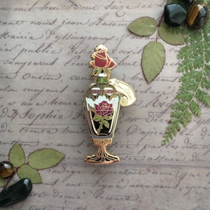 May include: Gold-tone enamel pin of a glass bottle with a rose inside. The bottle has a gold stopper and a tag that says "Potion".