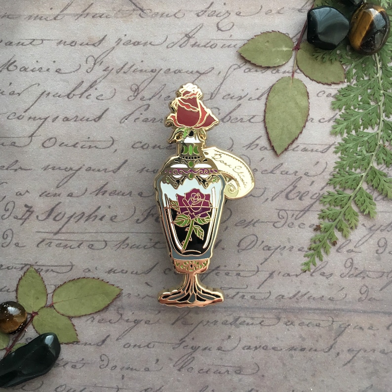 May include: Gold-tone enamel pin of a glass bottle with a rose inside. The bottle has a gold stopper and a tag that says "Potion".