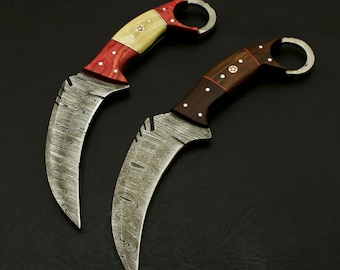 Può includere: Due coltelli curvi con lame in acciaio Damasco e manici in legno. I manici sono decorati con legno rosso e bianco su un coltello e legno marrone e rosso sull'altro. Entrambi i coltelli hanno un anello sul manico.
